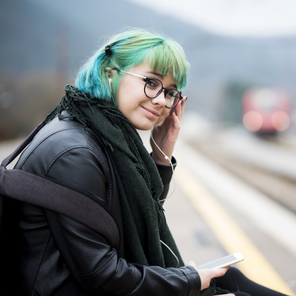 A content looking teenager with blue green hair sits by a train track