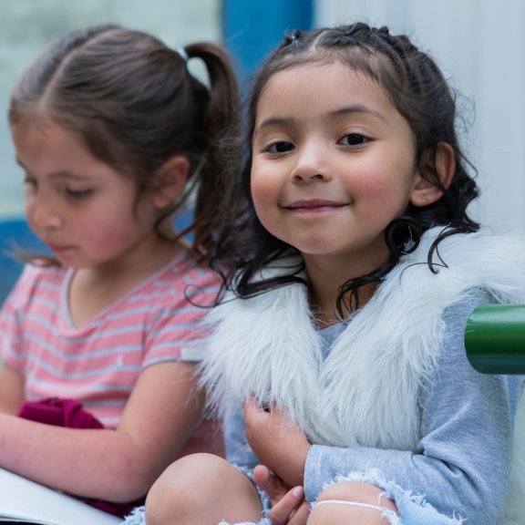 Two young girls sit next to each other. One looks at her book, the other smiles at the camera.