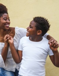 Adult and child smile at each other. Adult is standing behind with arms on either side of the child holding hands