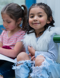 Two young girls sit next to each other. One looks at her book, the other smiles at the camera.